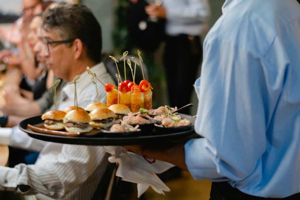 A waiter serves gourmet appetizers, including mini burgers, to patrons inside a bustling restaurant.