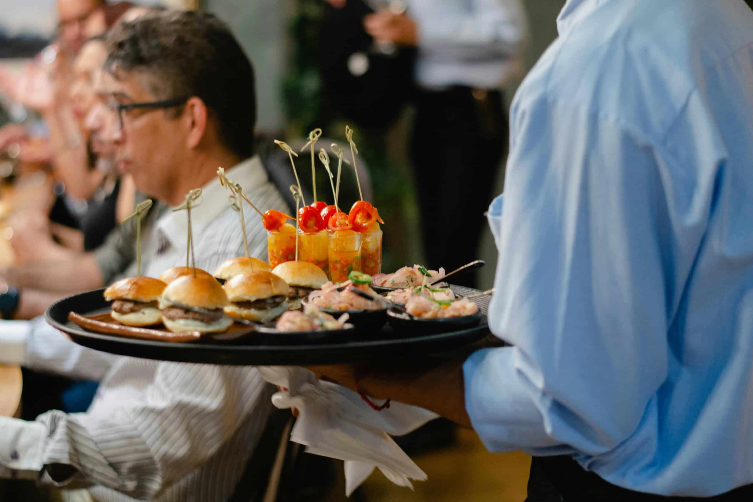 A waiter serves gourmet appetizers, including mini burgers, to patrons inside a bustling restaurant.